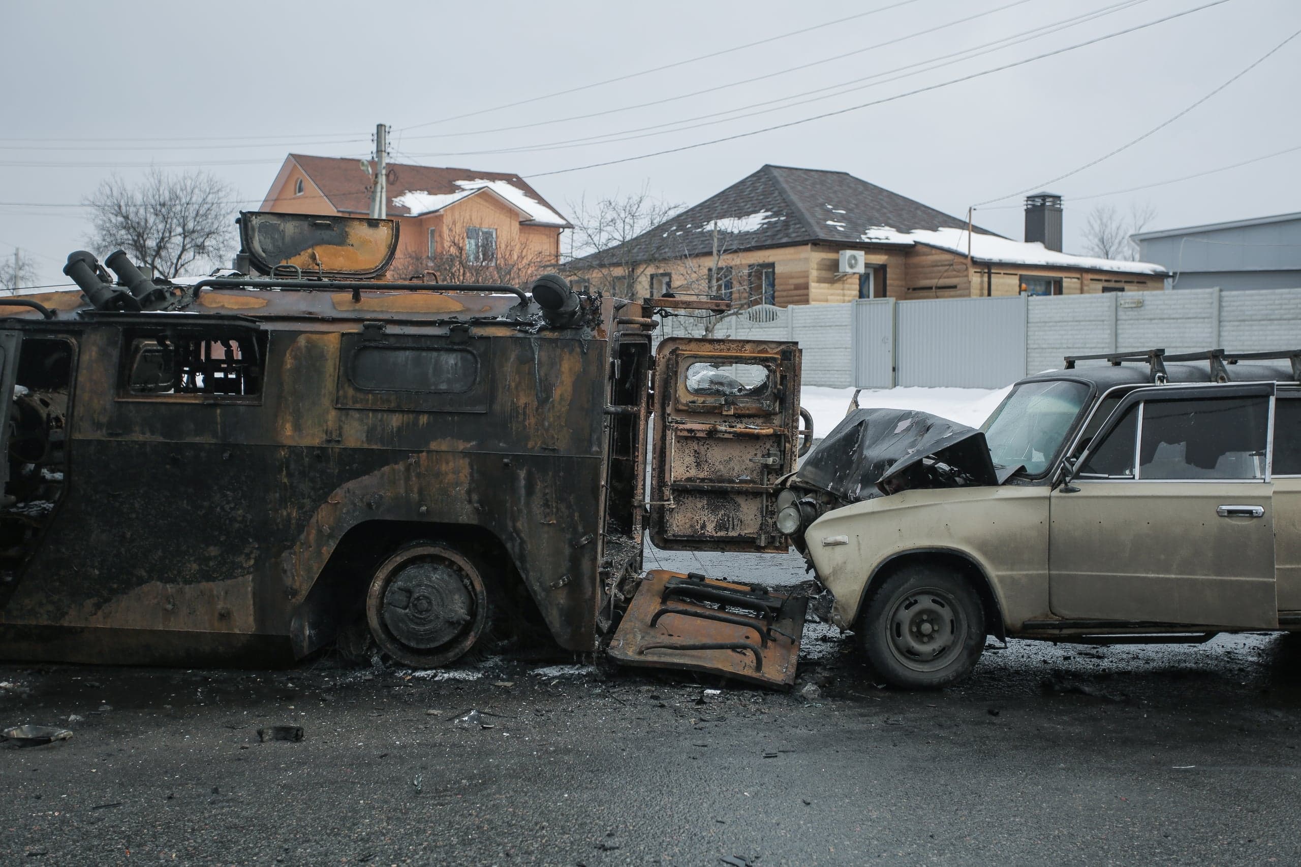 Сгоревший бронетранспортер, столкнувшийся с поврежденным легковым автомобилем, на фоне жилых домов.