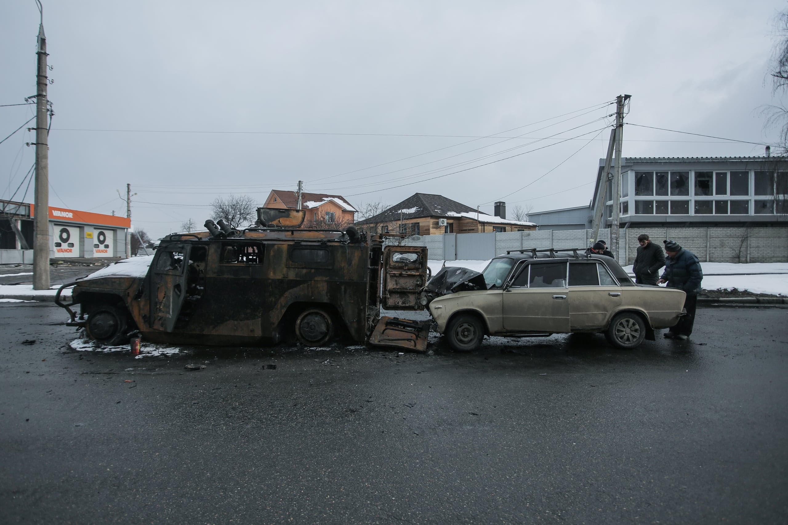 Сгоревший бронеавтомобиль и поврежденный легковой автомобиль на дороге, рядом двое мужчин. На фоне жилые дома и коммерческое здание с надписью "VARDE".