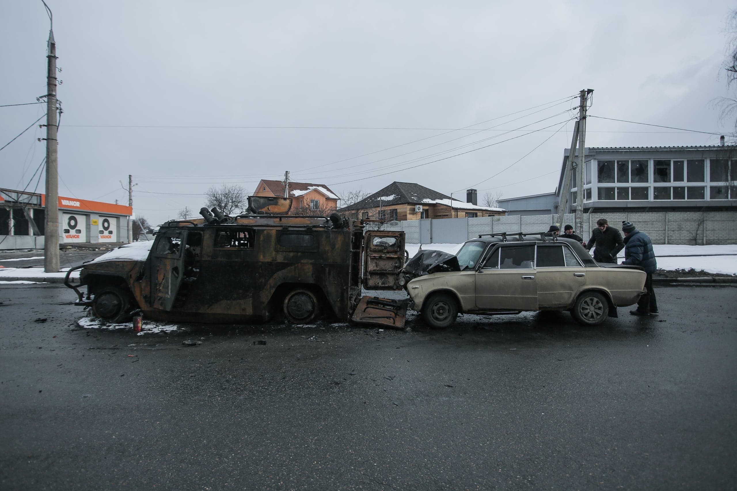 Сгоревший бронеавтомобиль и поврежденный легковой автомобиль после ДТП или боевых действий на улице, рядом несколько человек.