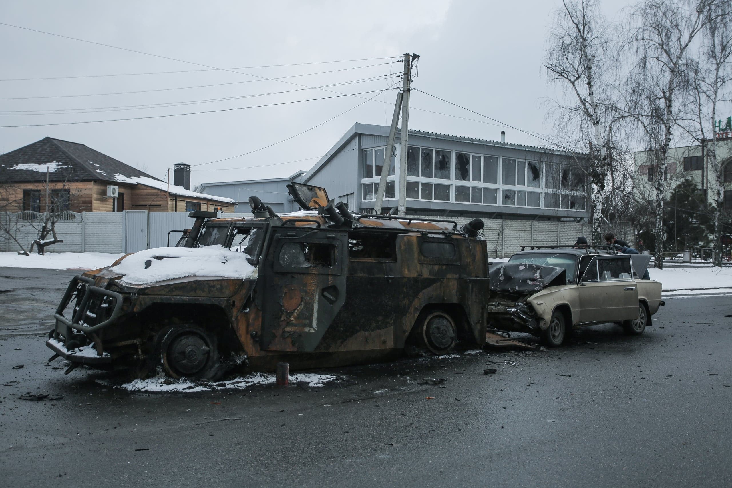 Сгоревший бронеавтомобиль и разбитый легковой автомобиль на заснеженной улице, на фоне зданий с выбитыми окнами.