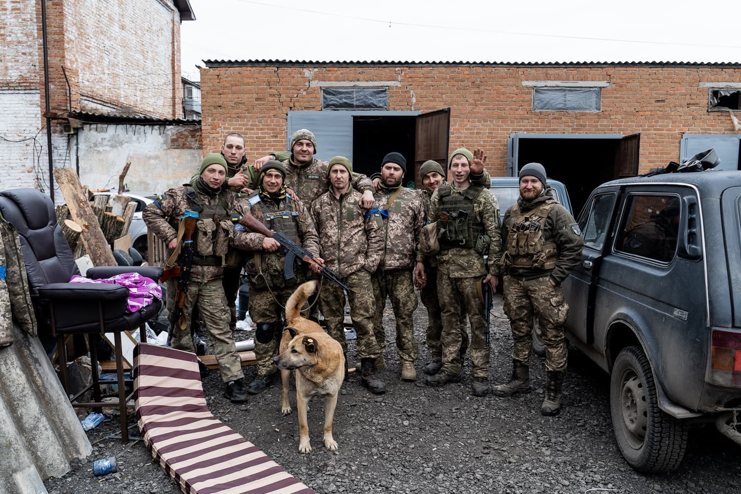 Группа из девяти солдат в камуфляжной форме с оружием и собака позируют на фоне кирпичного здания и внедорожника.