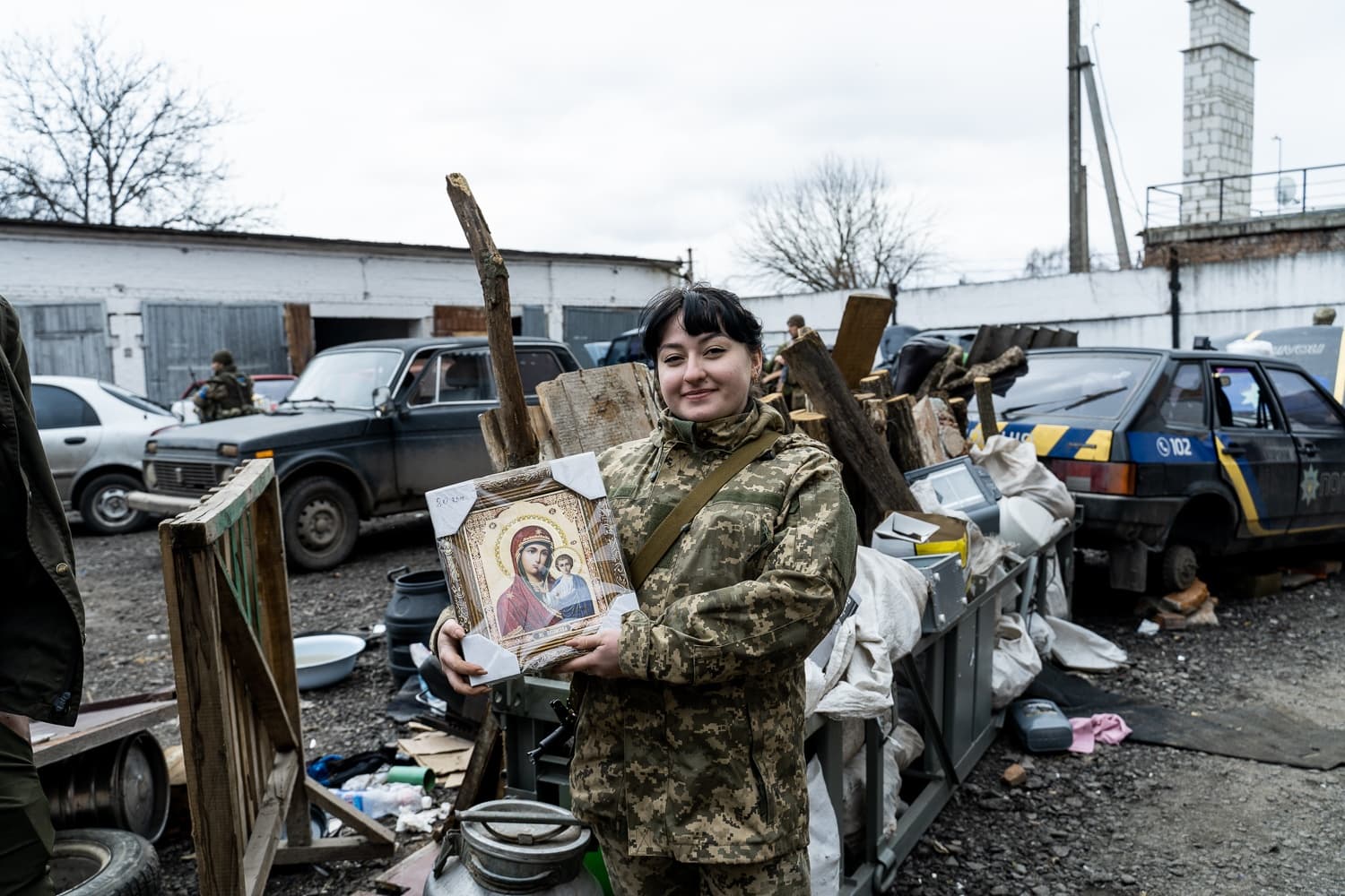 Улыбающаяся военнослужащая в камуфляже держит икону Богородицы с надписью "БОГОМАТІР" на фоне обломков и старых машин.