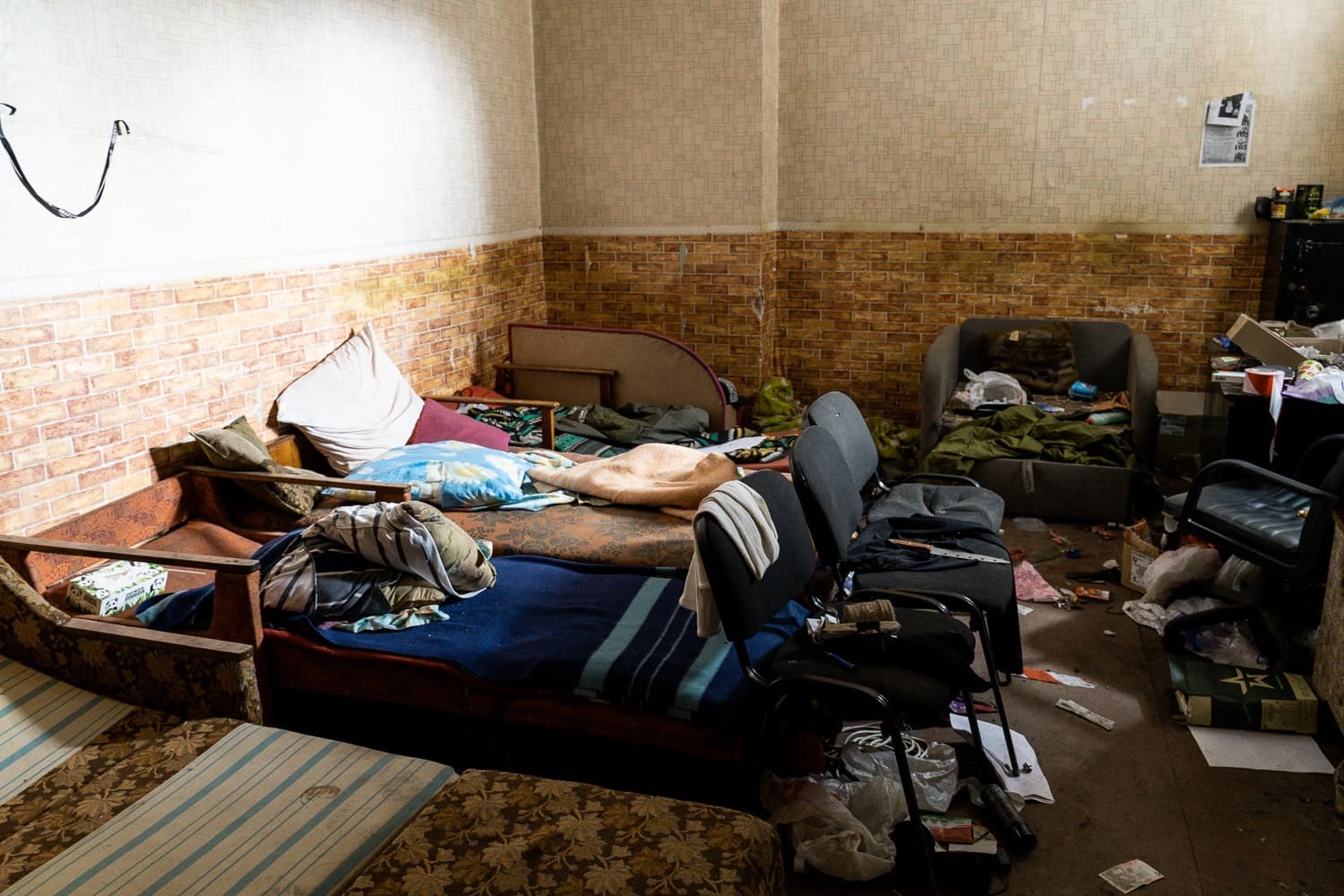 Грязное захламленное помещение с несколькими матрасами, используемыми как кровати, старыми обоями и мусором на полу.