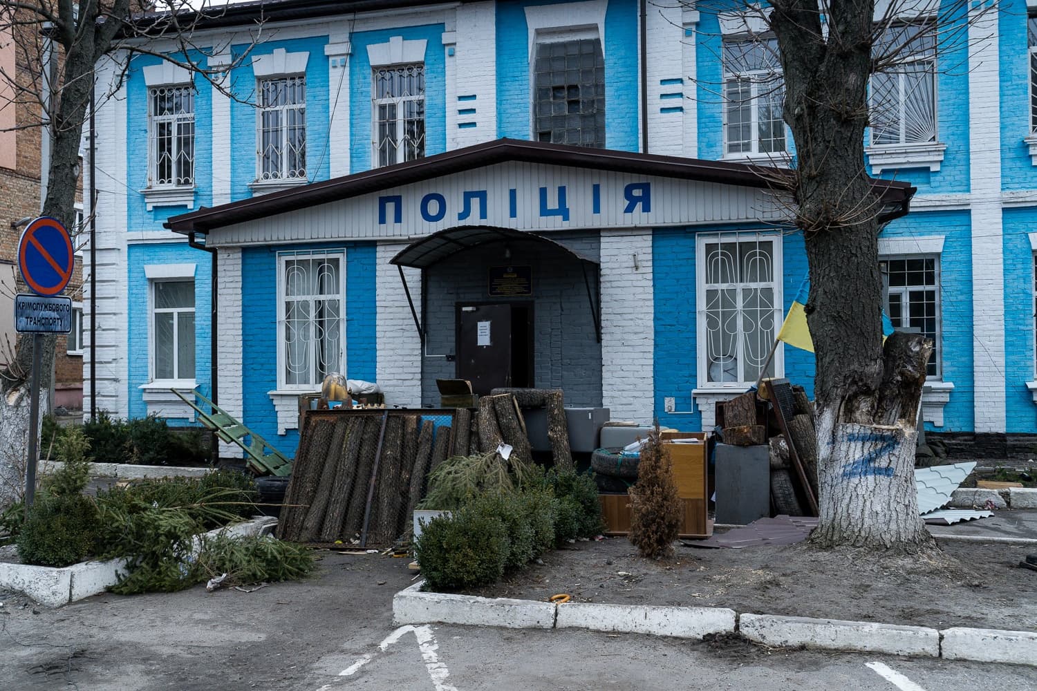 Голубое и белое здание полиции с вывеской "ПОЛІЦІЯ". Перед входом навалены бревна, обломки и мусор. На стволе дерева справа нарисована синяя буква Z и виден украинский флаг.