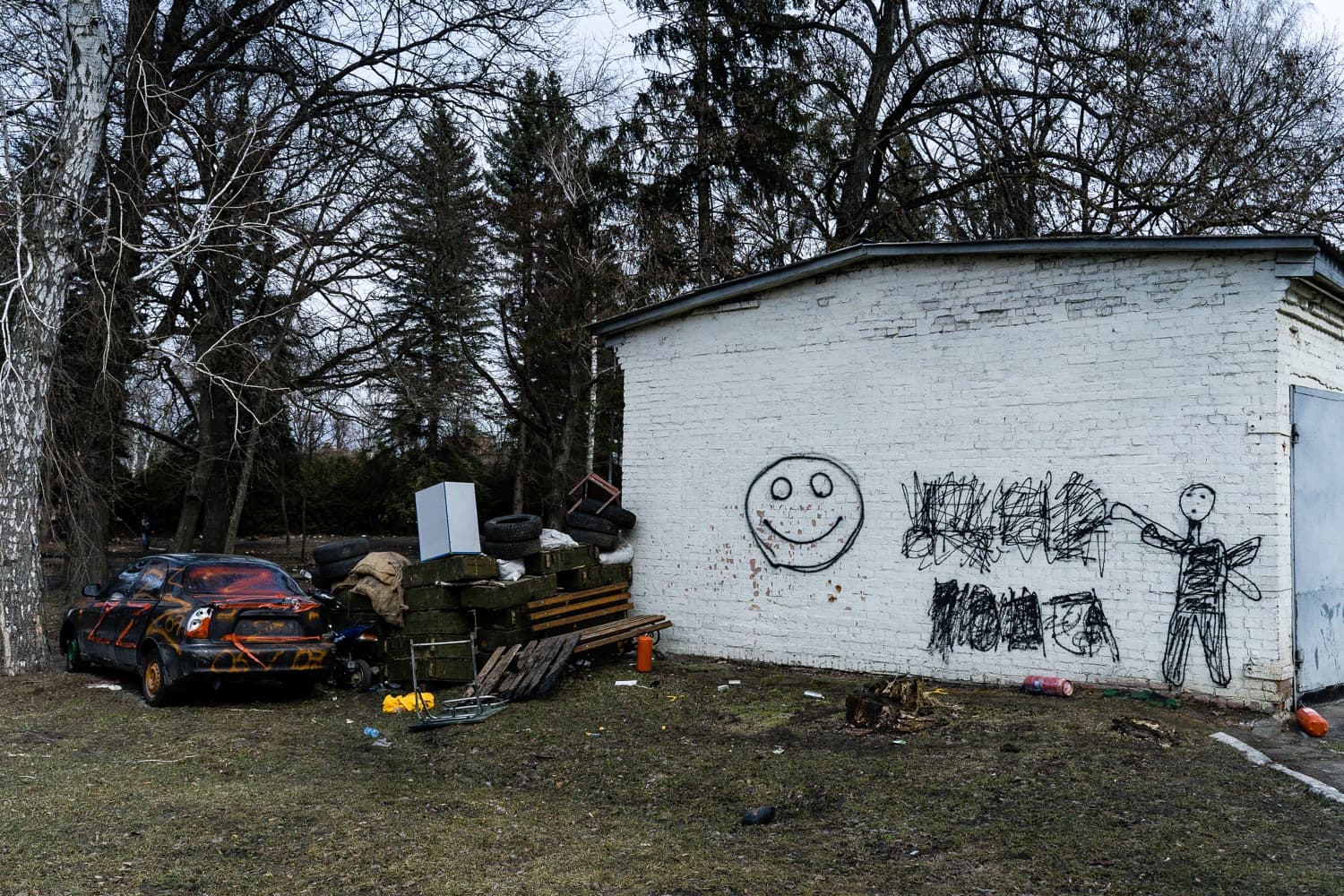 Обгоревшая машина и куча мусора на фоне белого кирпичного сарая. На стене нарисованы граффити: большой смайлик и стилизованный человечек.