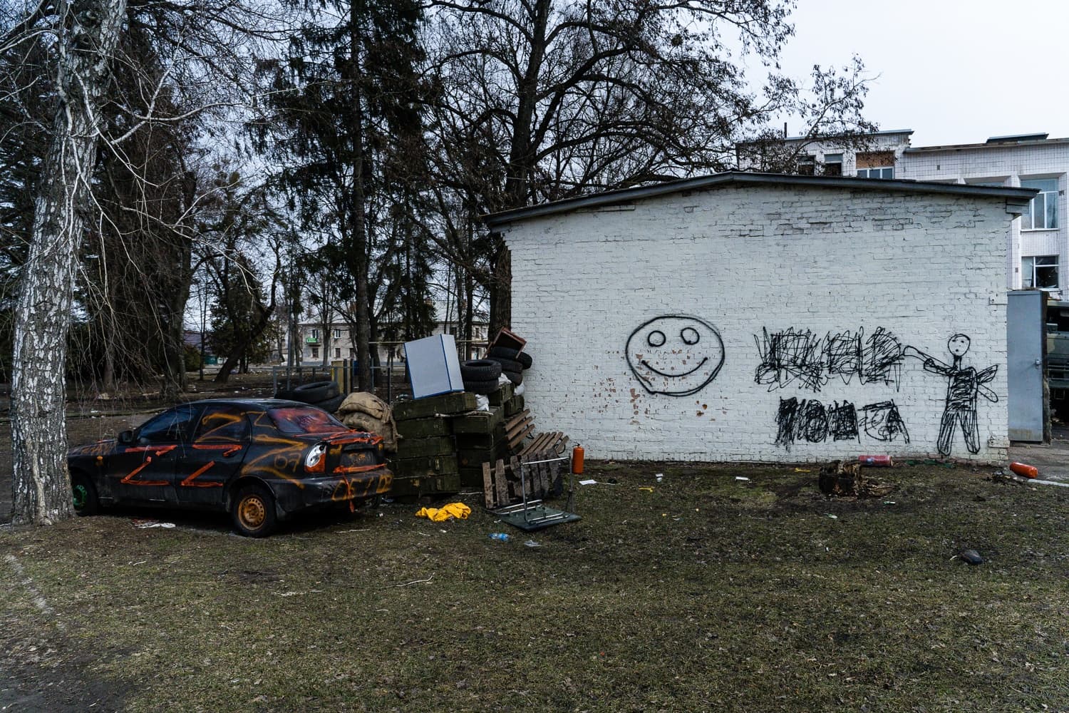 Разрушенный черный автомобиль с оранжевыми символами "Z" стоит у белой стены с граффити (смайлик и фигура человека) на фоне груды обломков.
