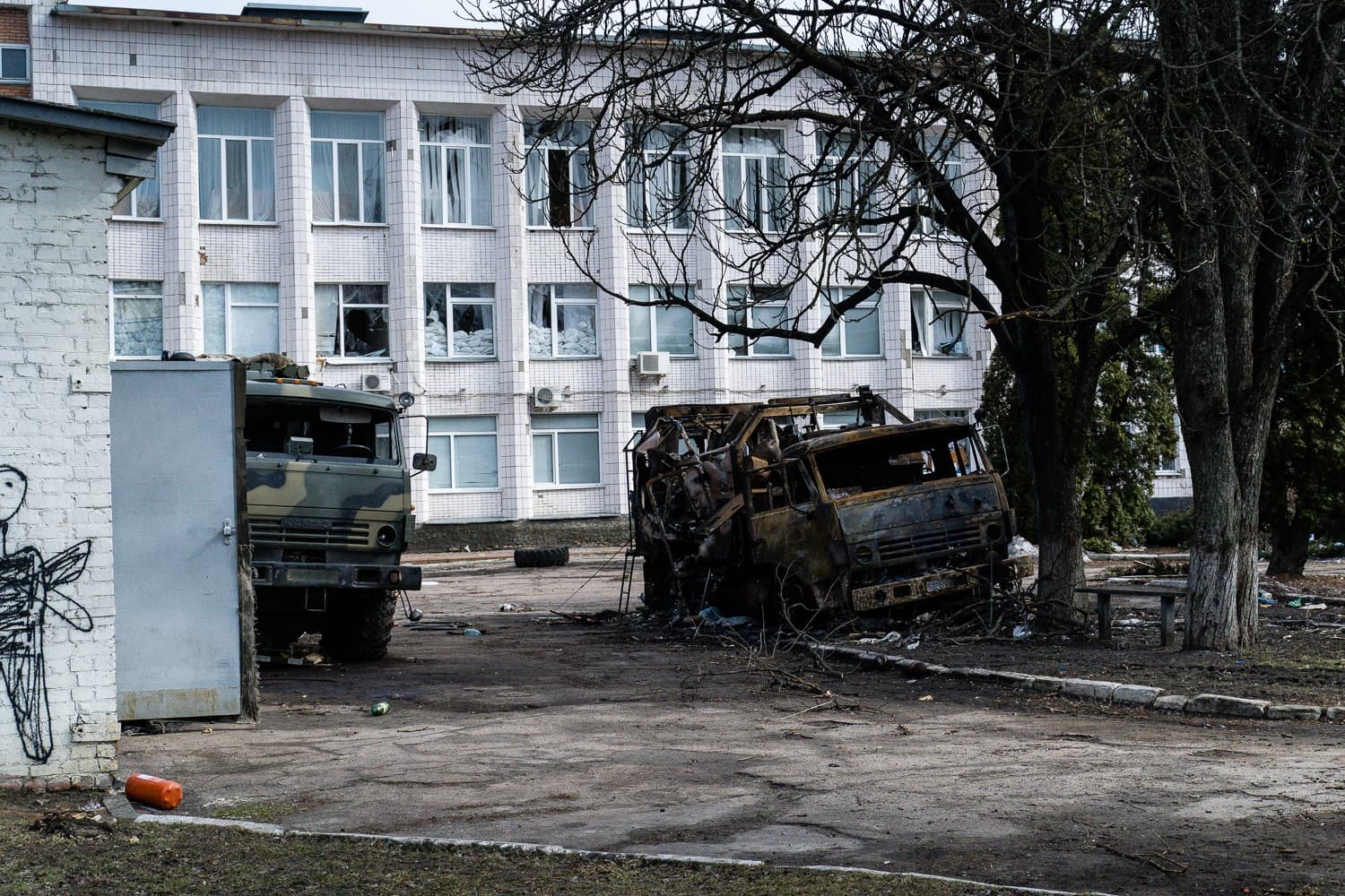 Сгоревший военный грузовик и камуфляжная машина перед зданием с разбитыми окнами.