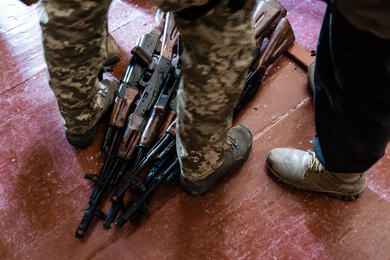 Ноги двух человек (один в камуфляже, другой в темных брюках) стоят над кучей автоматов Калашникова на красном полу.