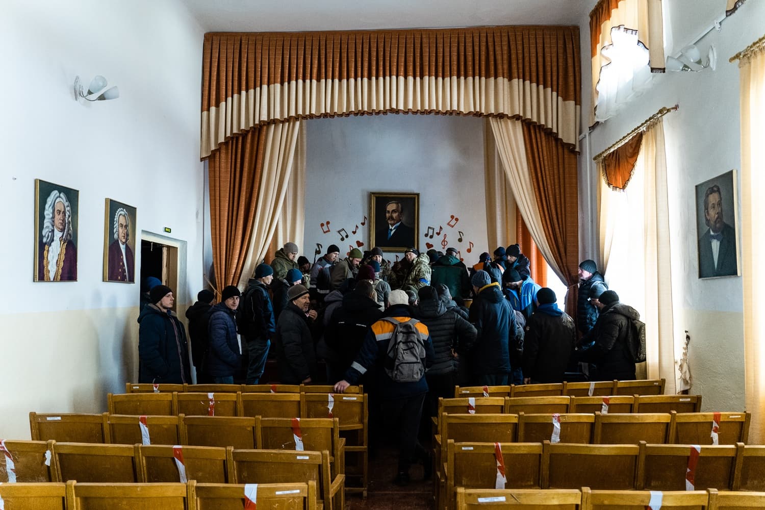 Актовый зал с пустыми деревянными стульями. Большая группа мужчин в зимней одежде собралась перед сценой, украшенной портретом и нотами.