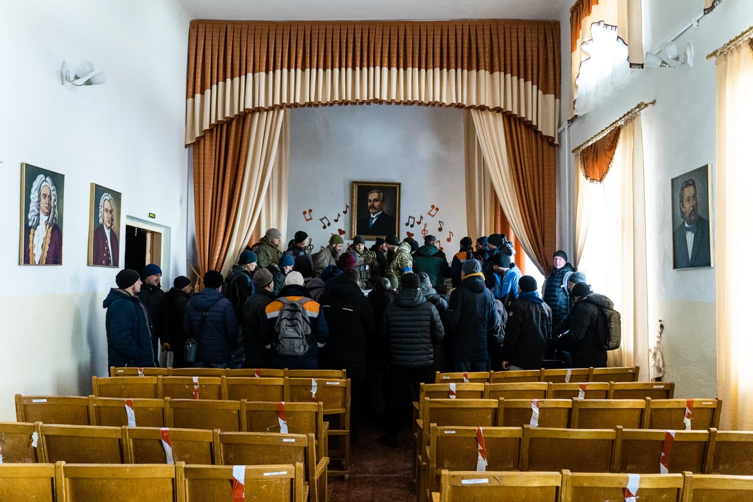 Группа людей в зимней одежде собралась у сцены актового зала с портретами и занавесами. На заднем плане висит портрет мужчины в центре, украшенный нотами. Ряды деревянных стульев в зале частично перевязаны красной и белой лентой.