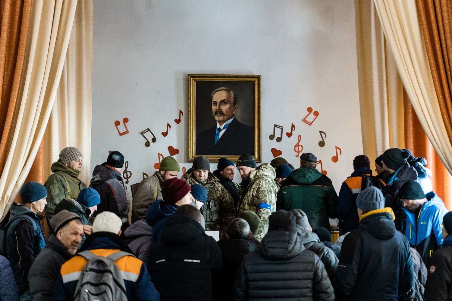 Толпа мужчин в зимней одежде в помещении. На стене висит портрет мужчины с усами, окруженный вырезанными музыкальными нотами.