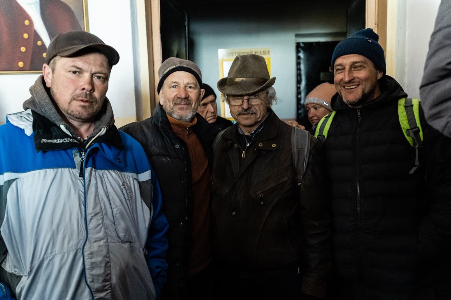 Группа из четырех мужчин в теплой одежде (куртки, шапки) и шляпе стоит в помещении. Мужчина справа широко улыбается.