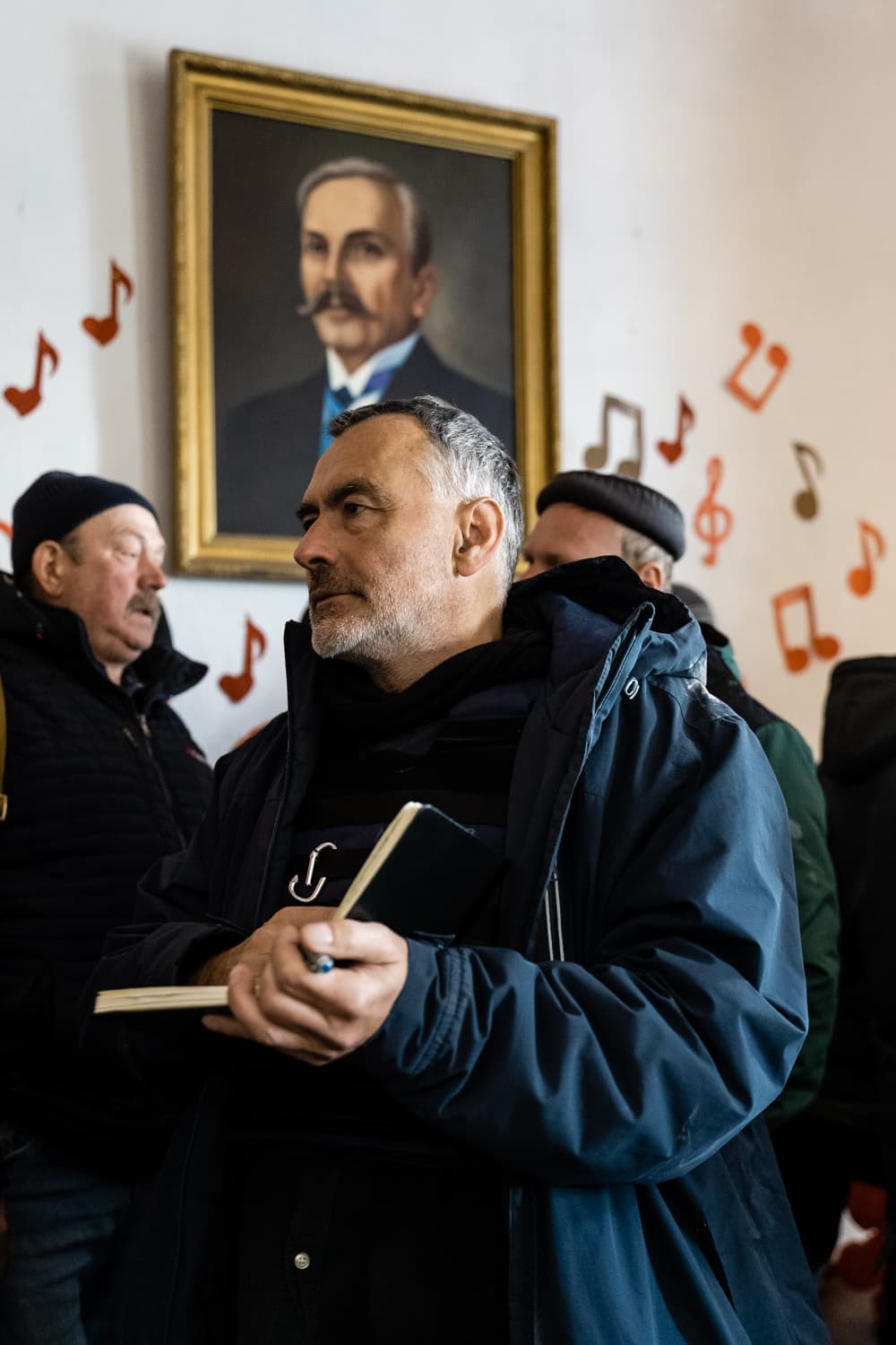 Мужчина средних лет в тёмной куртке держит блокнот и ручку на фоне портрета в золотой раме и стилизованных нот на стене.