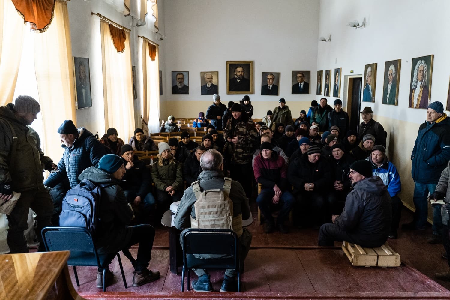 Группа людей слушает инструктора в тактическом жилете в лекционной аудитории. Стены помещения украшены портретами.