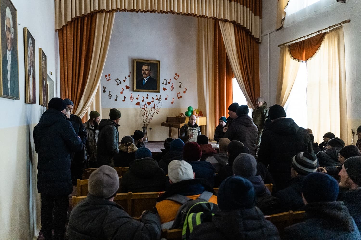 Собрание людей в зимней одежде в актовом зале. На стене за сценой висит портрет, украшенный вырезками нот и красных сердец. Оратор обращается к сидящей аудитории.