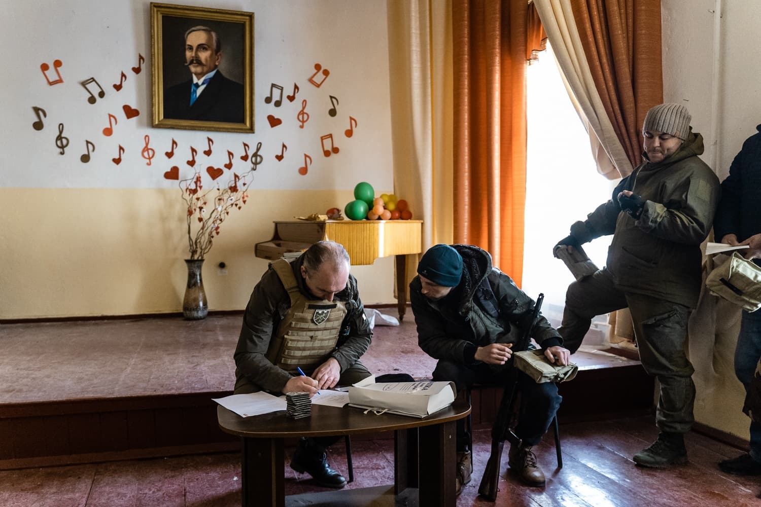 Двое военных в бронежилетах заполняют бумаги за столом на сцене, украшенной нотами, сердцами и портретом.