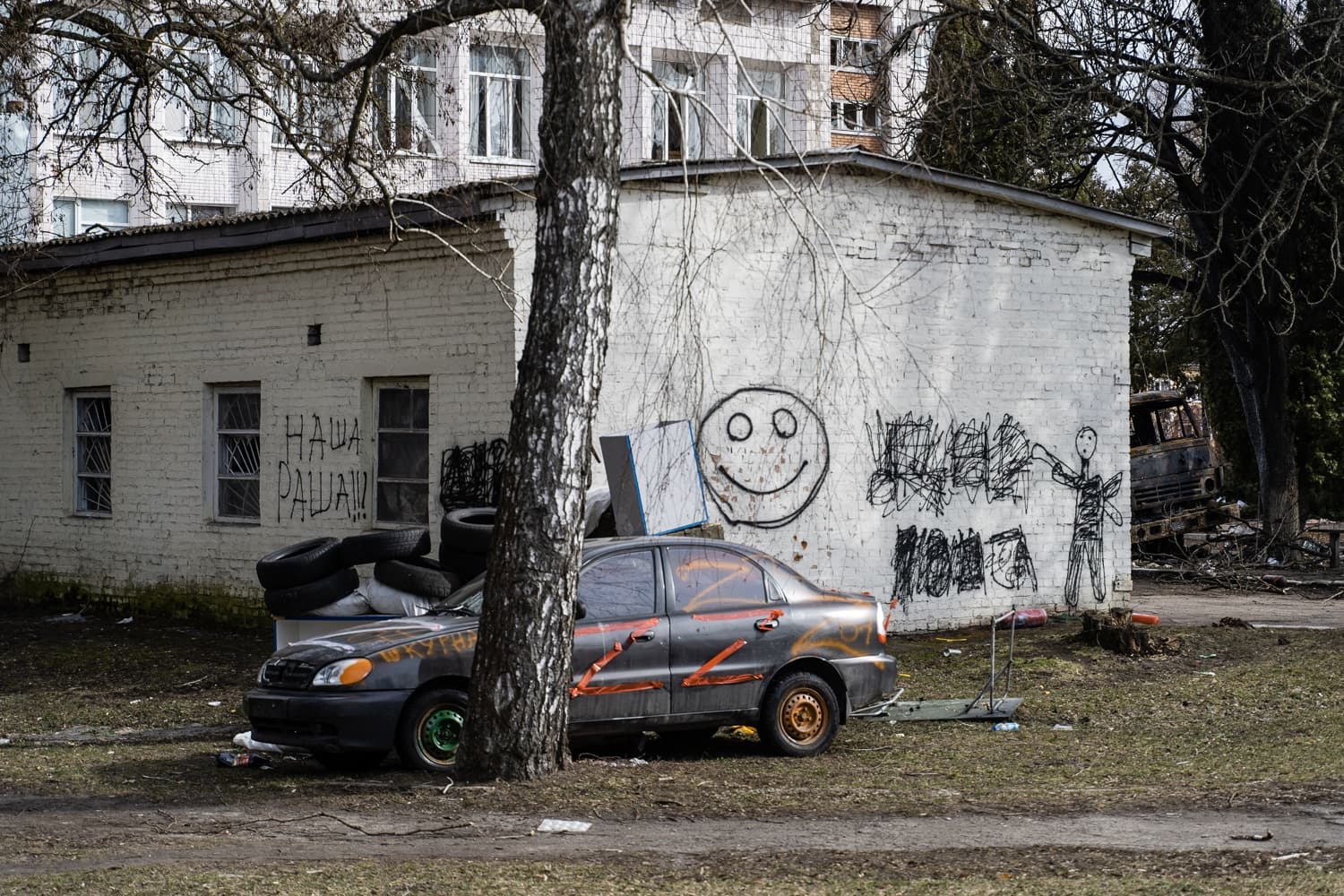Разрисованный автомобиль с символами Z на фоне белого здания с граффити, включая "НАША РАША!!!" и смайлик.