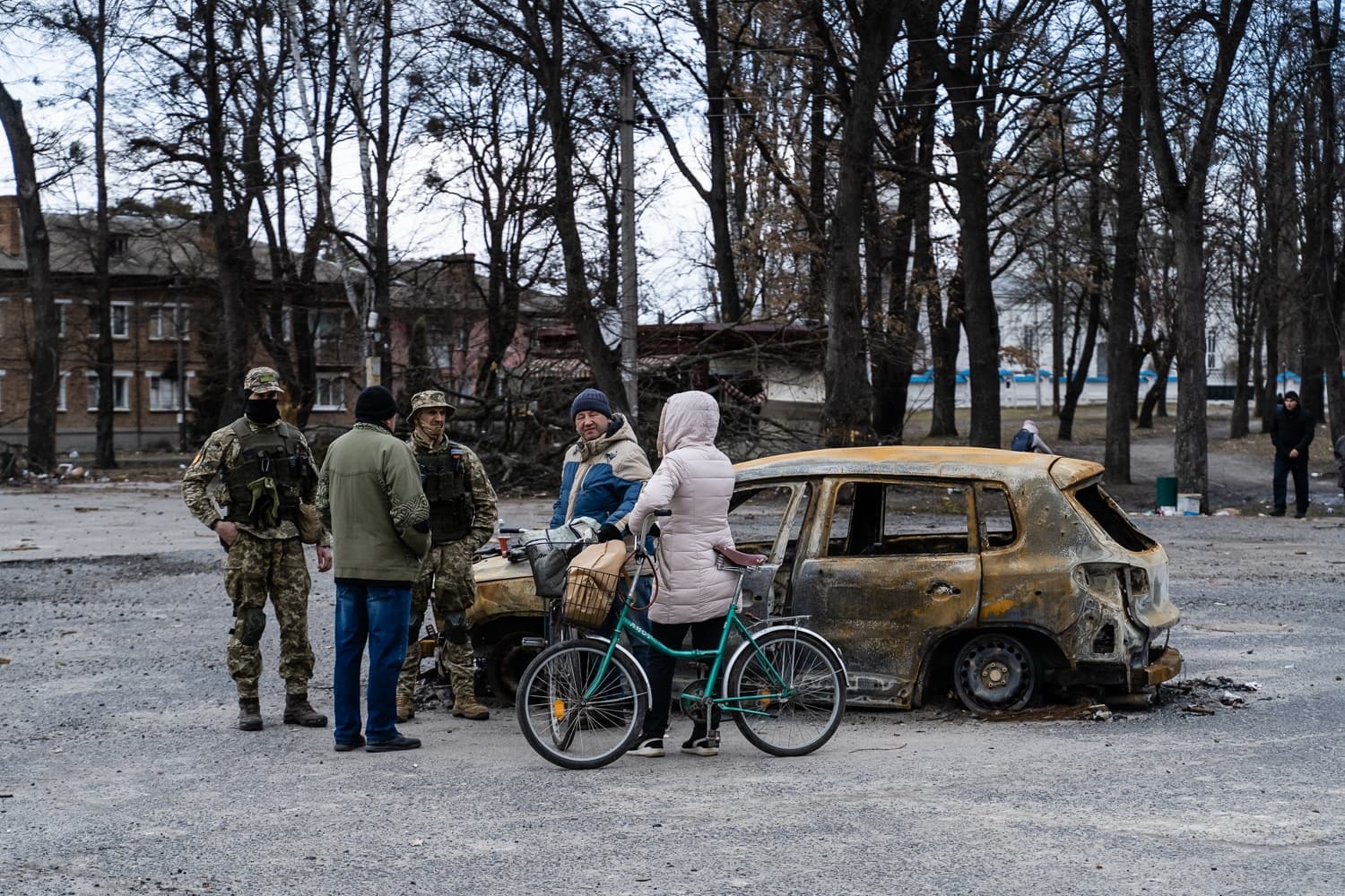 Два солдата и трое гражданских стоят рядом со сгоревшим автомобилем на разрушенной улице. Женщина держит велосипед.