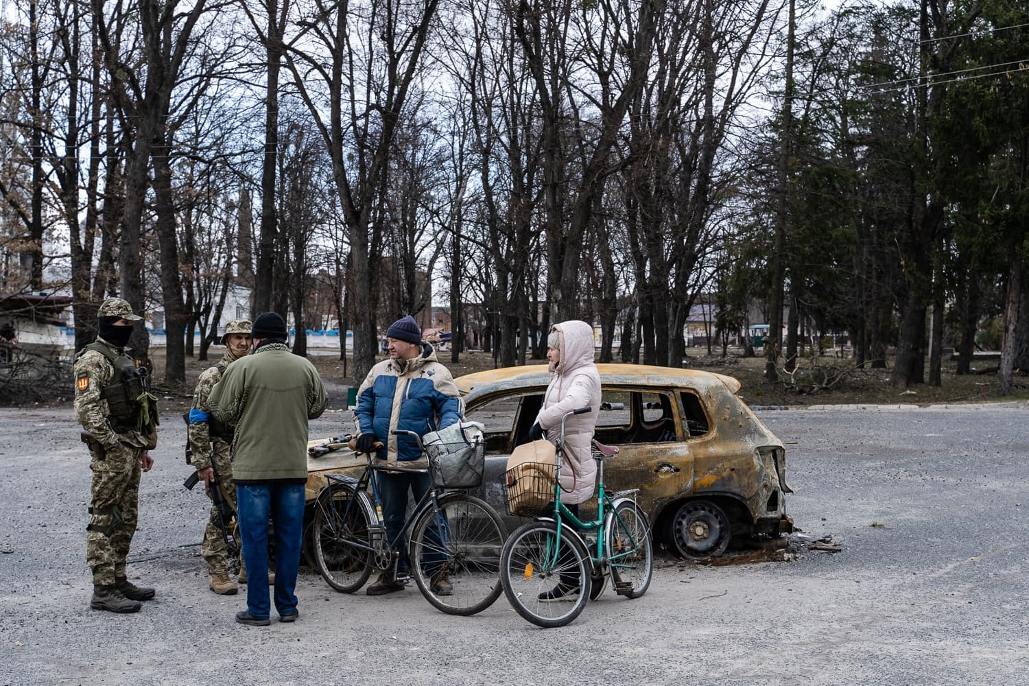 Гражданские с велосипедами разговаривают с военными рядом со сгоревшим автомобилем на фоне голых деревьев.