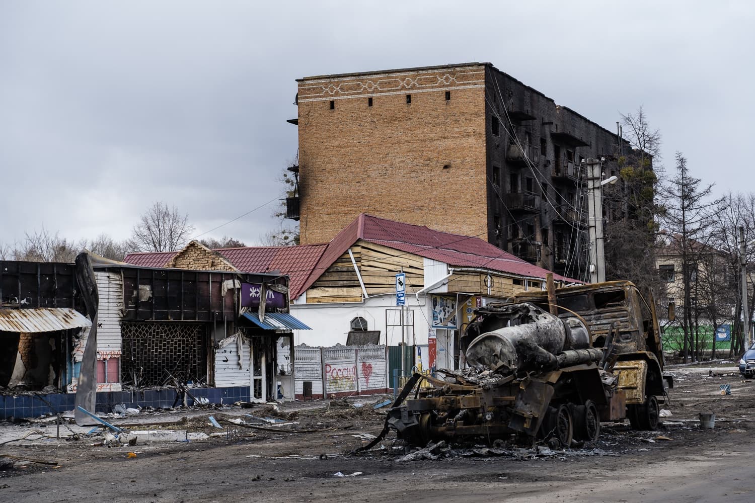 Обугленный грузовик-цистерна перед сильно поврежденными и выгоревшими зданиями. Задний фасад многоэтажного кирпичного дома полностью почернел от огня. На заборе граффити «РОССИЯ».