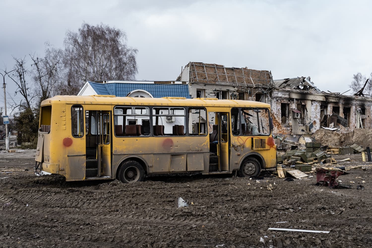 Поврежденный желтый автобус в грязи на фоне разрушенных войной зданий.