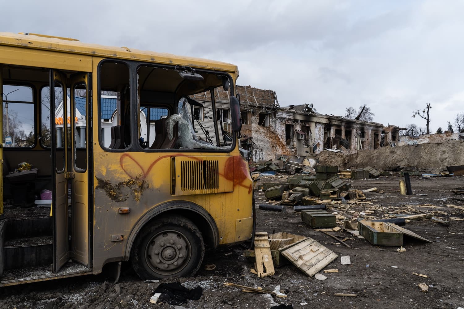 Разрушенный желтый автобус на фоне руин сильно поврежденного кирпичного здания. На земле разбросаны обломки и зеленые военные ящики.