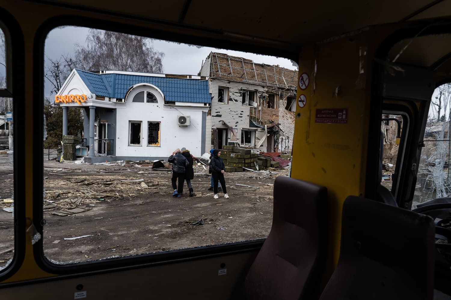 Вид из автобуса на разрушенные войной здания, засыпанную мусором улицу и несколько стоящих людей. На уцелевшем здании вывеска: "Дніпро".