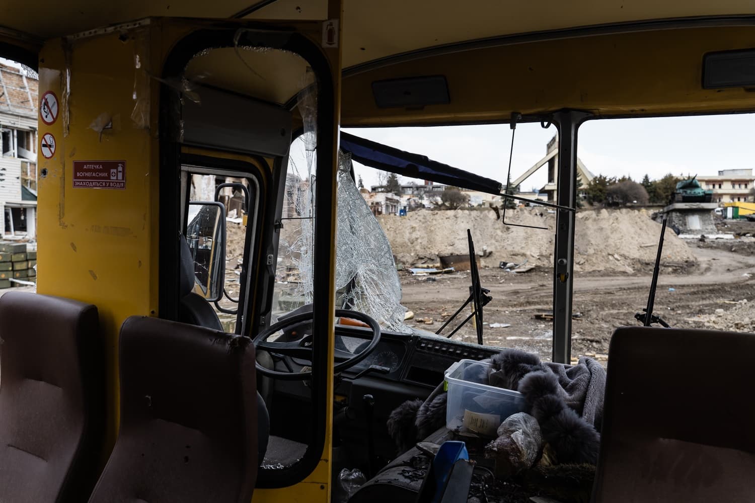 Салон поврежденного желтого автобуса с разбитым лобовым стеклом. Вид на разрушенный пейзаж с насыпями и военной техникой. Текст: "АПТЕЧКА ВОГНЕТАСНИК ЗНАХОДЯТЬСЯ У ВОДІЯ".