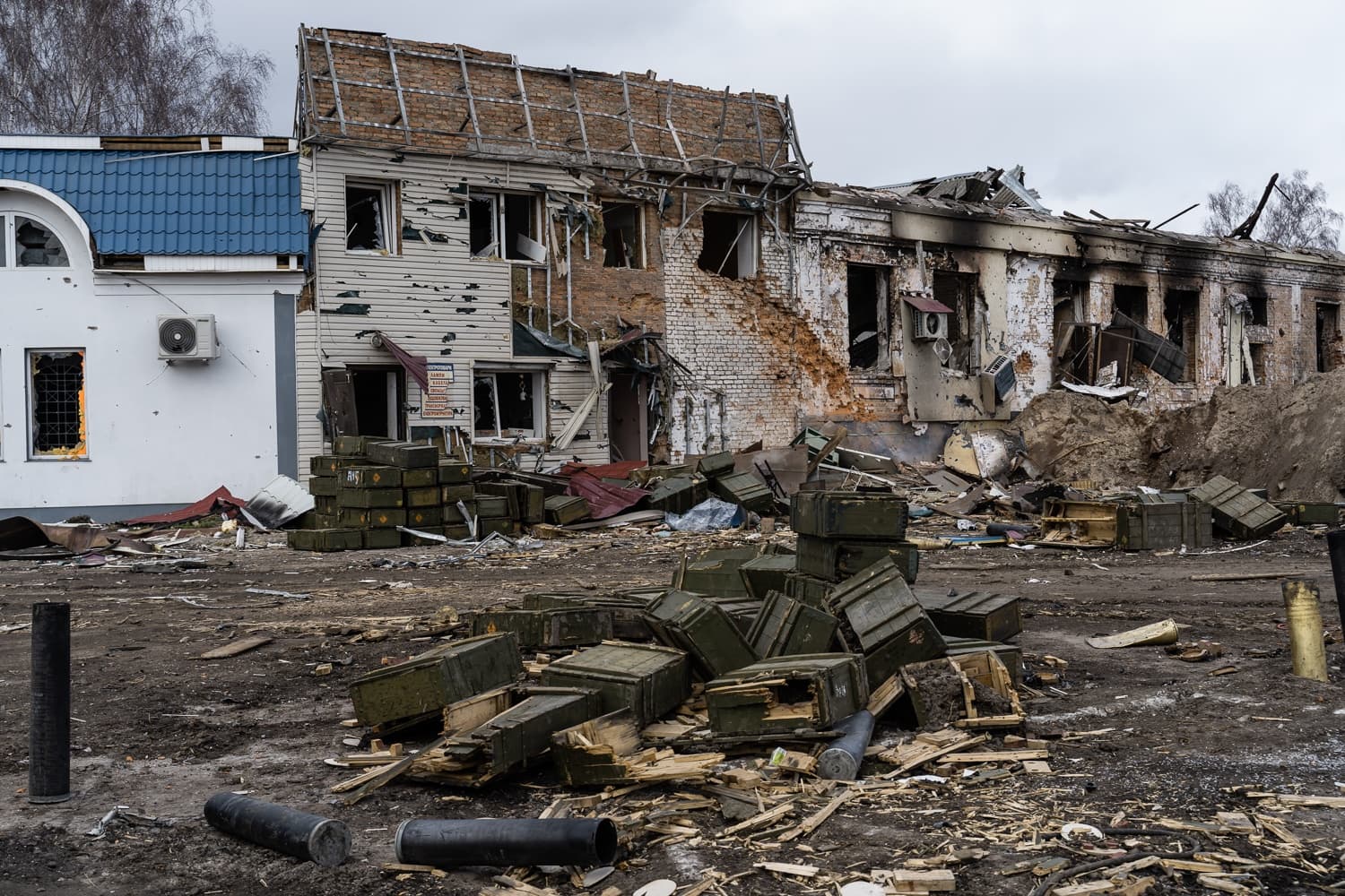Разрушенное здание с дырами от снарядов и обвалившейся крышей; на переднем плане лежат груды деревянных ящиков для боеприпасов и крупные гильзы.