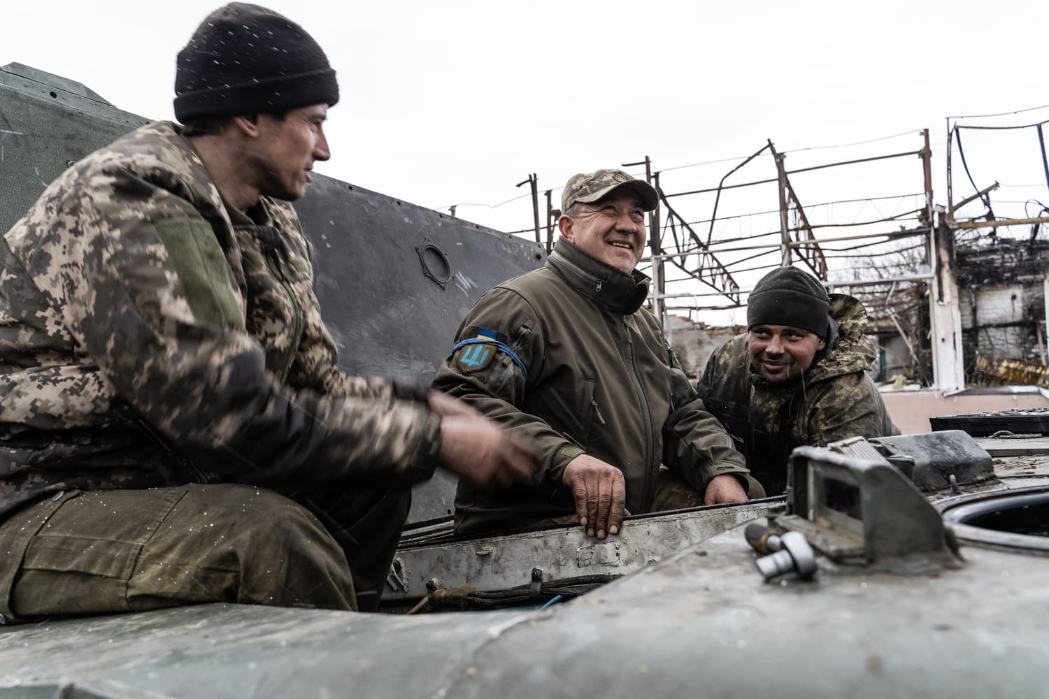Трое украинских военнослужащих улыбаются, находясь в люке бронемашины на фоне разрушенных строений.
