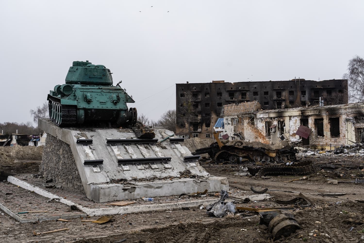 Памятник танку Т-34 на фоне обгоревших и разрушенных городских зданий. На земле — обломки и остов сгоревшей техники.