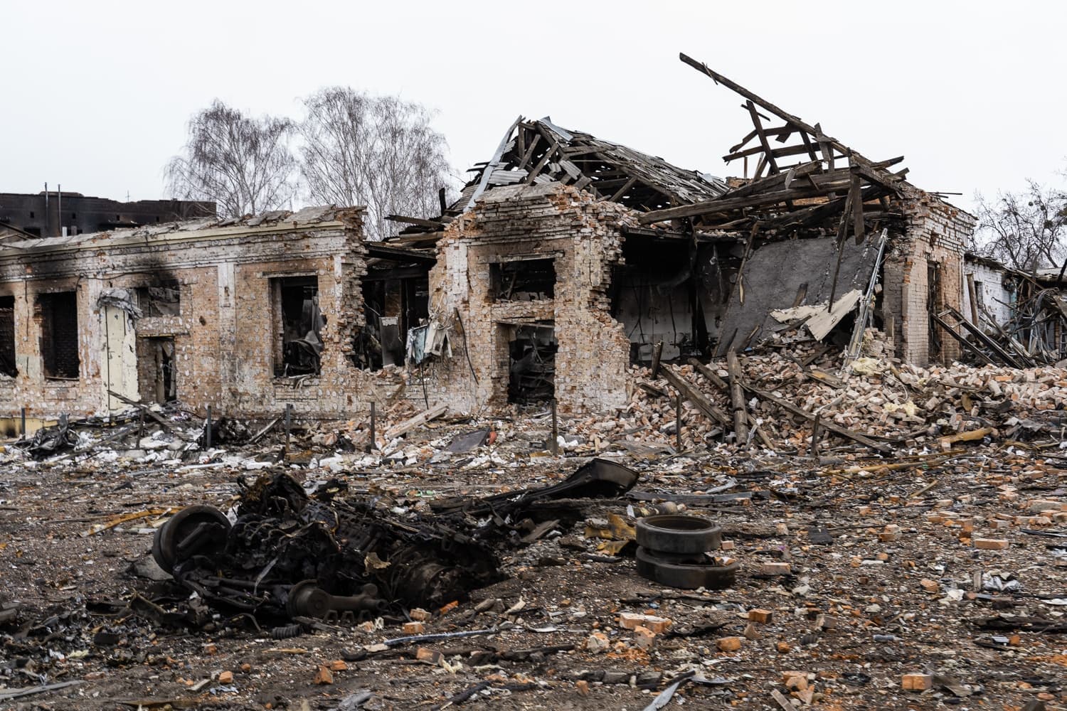 Разрушенное кирпичное здание с обрушенной крышей, окруженное грудами обломков и сгоревших деталей на переднем плане.