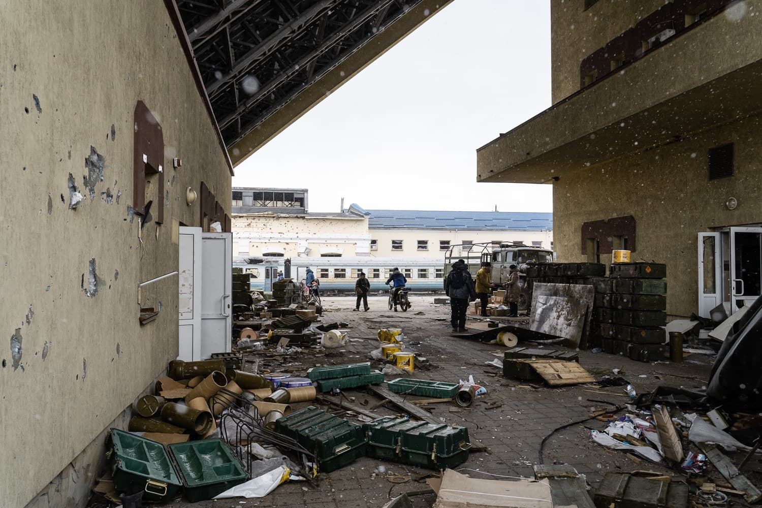 Разрушенный перрон вокзала, заваленный зелеными военными ящиками и гильзами. На стенах видны следы обстрела. На заднем плане люди идут мимо поезда и грузовика.