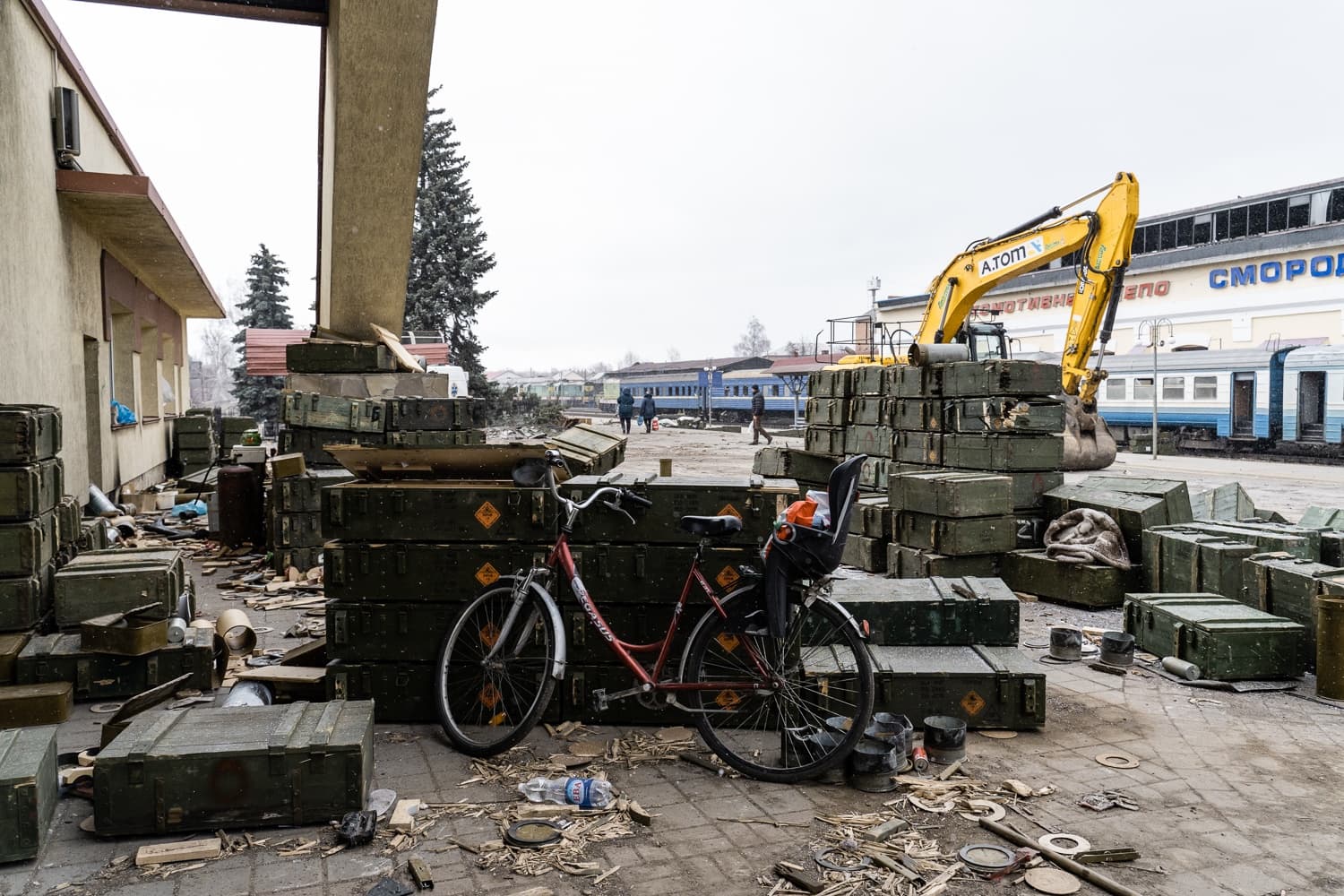 Велосипед с детским креслом среди штабелей зеленых военных ящиков на вокзале. Желтый экскаватор, на здании станции надпись "СМОРОД".