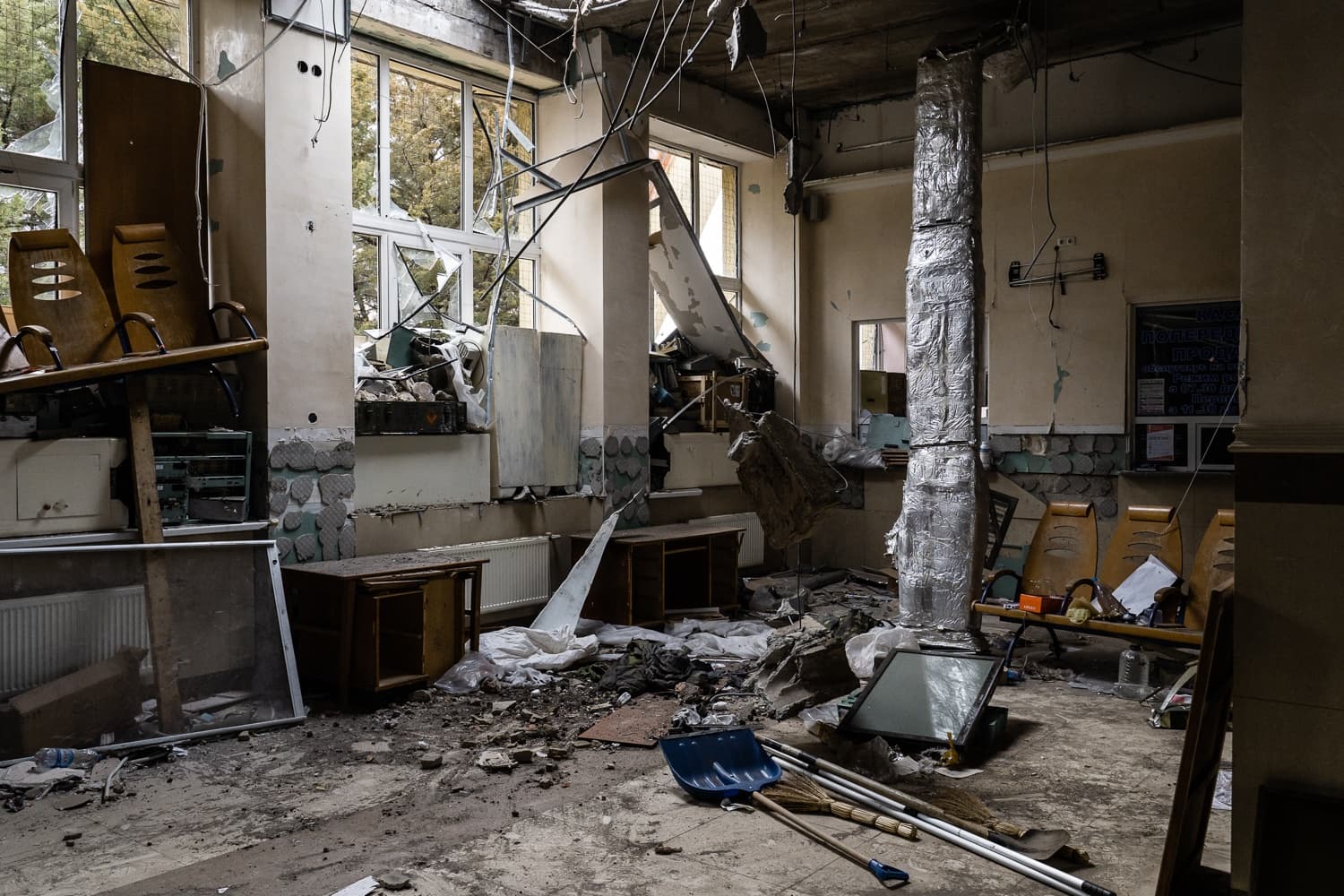 Сильно разрушенное помещение, заваленное обломками: разбитые окна, поваленная мебель, пыль и строительный мусор на полу.