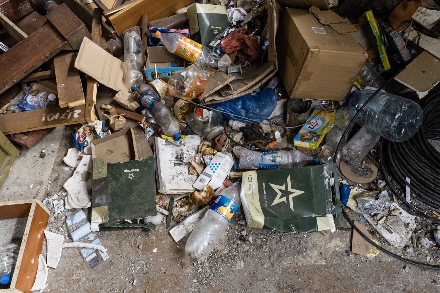 Куча мусора: пластиковые бутылки, картон, деревянные обломки и темно-зеленая упаковка с логотипом звезды и надписями "Армия России" и "Военторг".