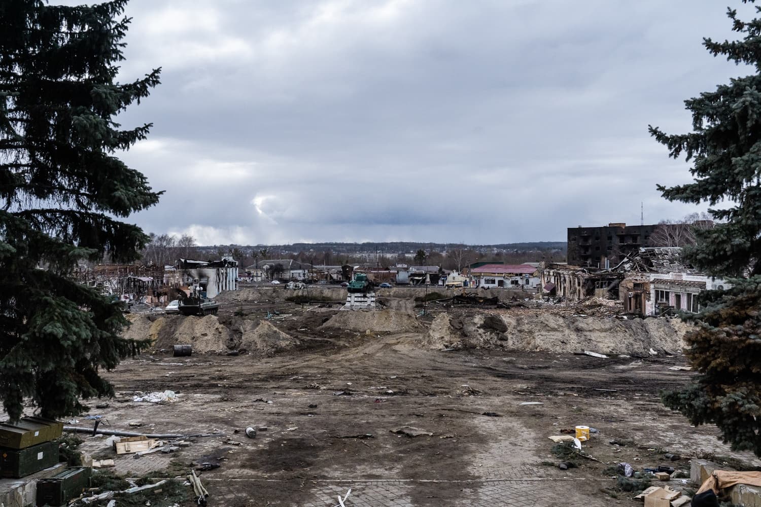Сильно разрушенная городская площадь, заполненная обломками и земляными насыпями, на фоне сгоревших зданий. Сцена обрамлена елями.