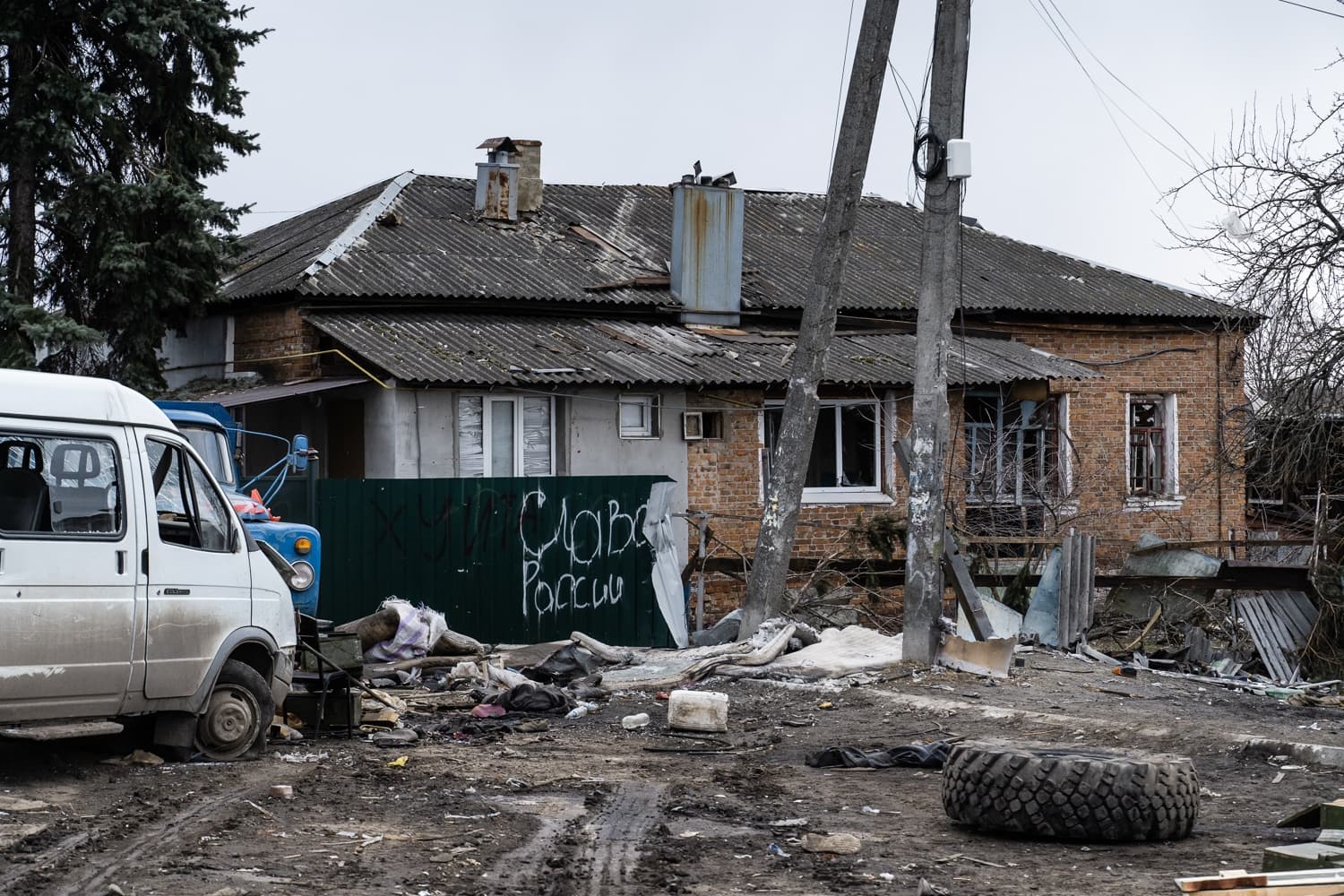 Разрушенный дом, грязный двор с обломками. На зеленом заборе белое граффити: "Слава России". Слева стоит поврежденный белый фургон.