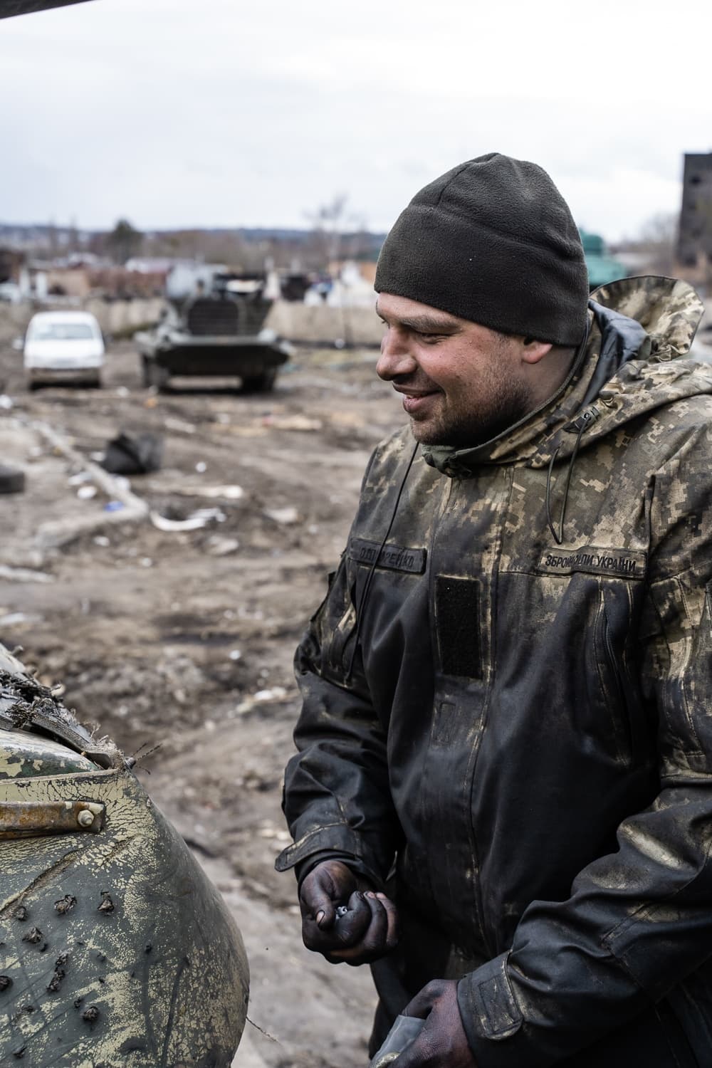 Грязный украинский солдат ("ЗБРОЙНІ СИЛИ УКРАЇНИ") улыбается, держа в руках металлический фрагмент, на фоне бронетехники и разрушенной местности.