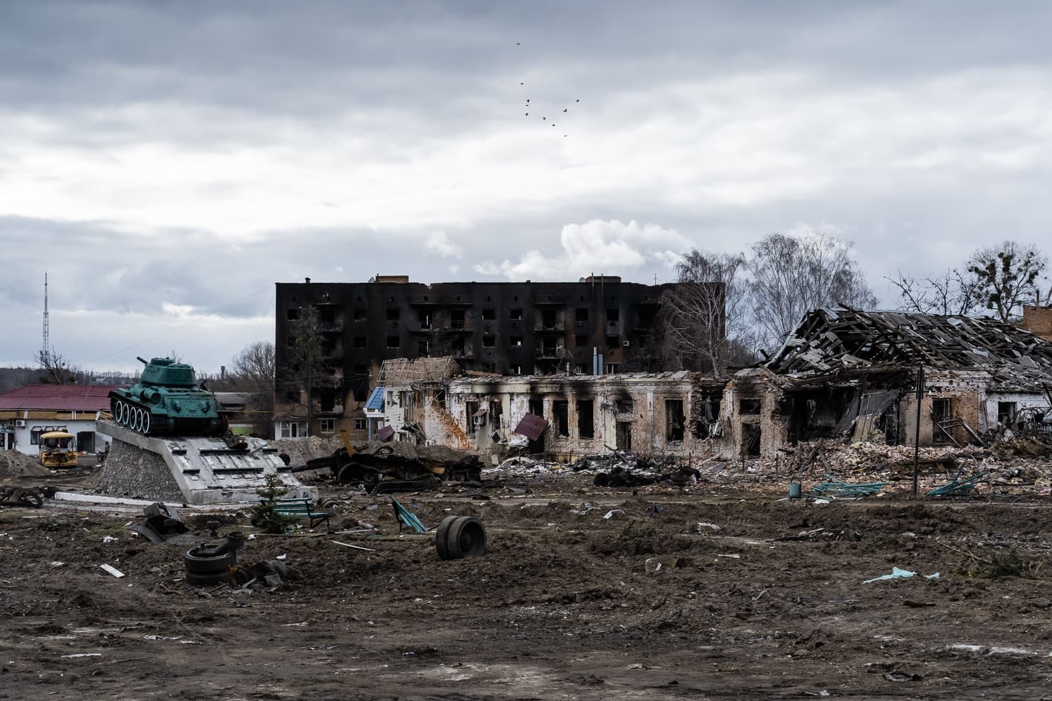 Танк-памятник на постаменте перед полностью разрушенными и обгоревшими многоквартирными домами.