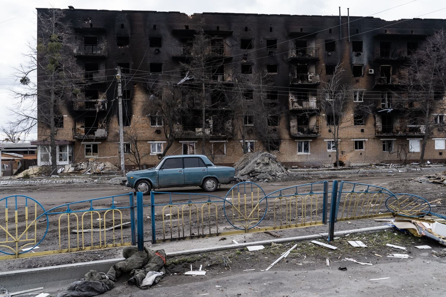 Сильно поврежденный огнем многоквартирный дом и разрушенная улица с припаркованным синим автомобилем и обломками.