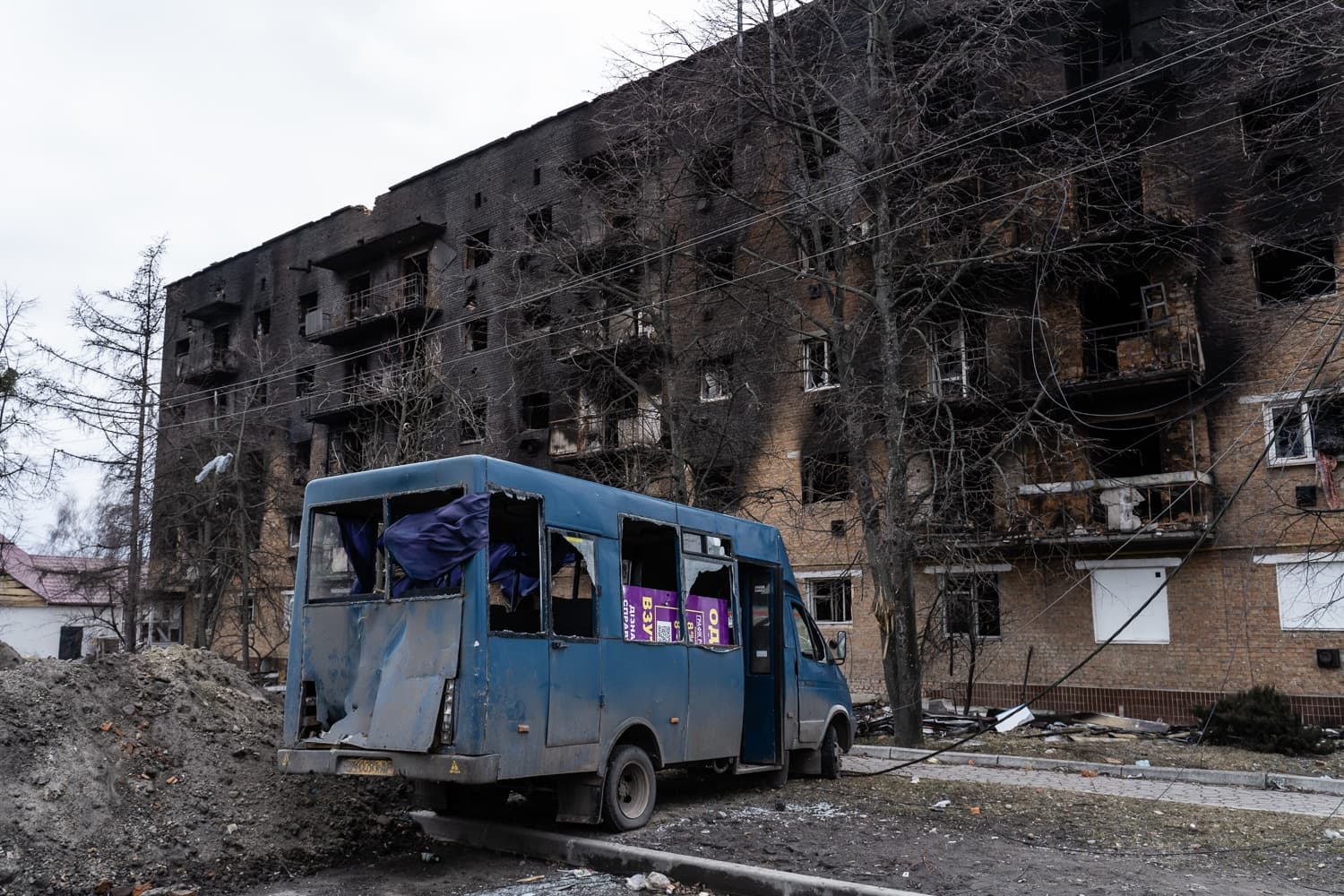 Поврежденный синий микроавтобус перед многоэтажным домом с полностью обгоревшим и почерневшим фасадом.
