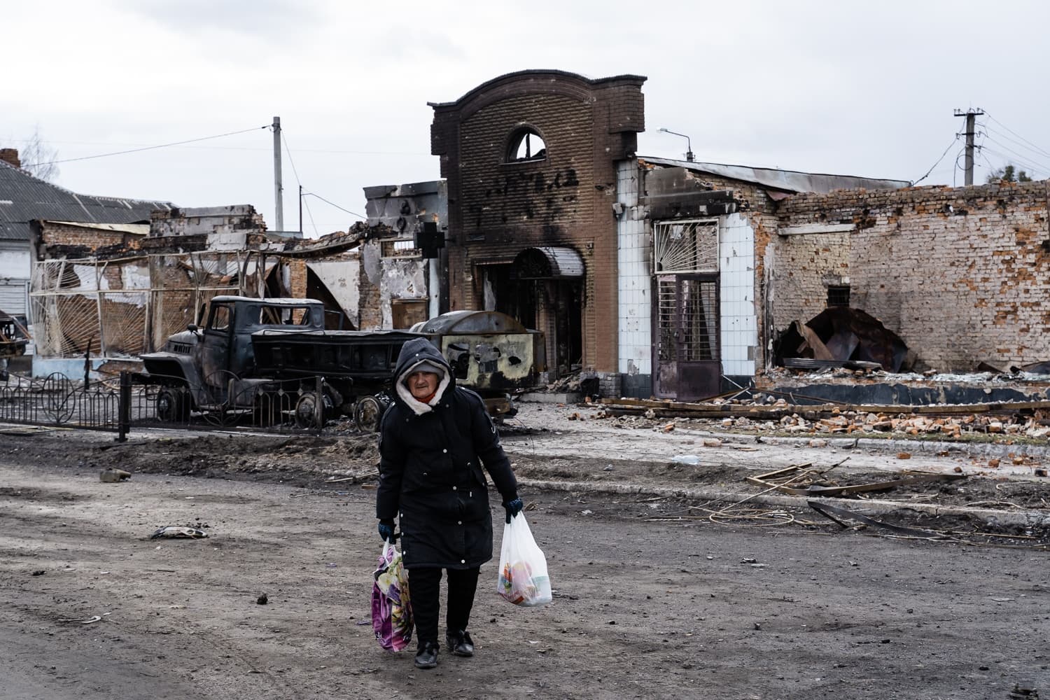 Пожилая женщина с сумками идет по улице на фоне разрушенных, обгоревших зданий и сгоревшего грузовика.