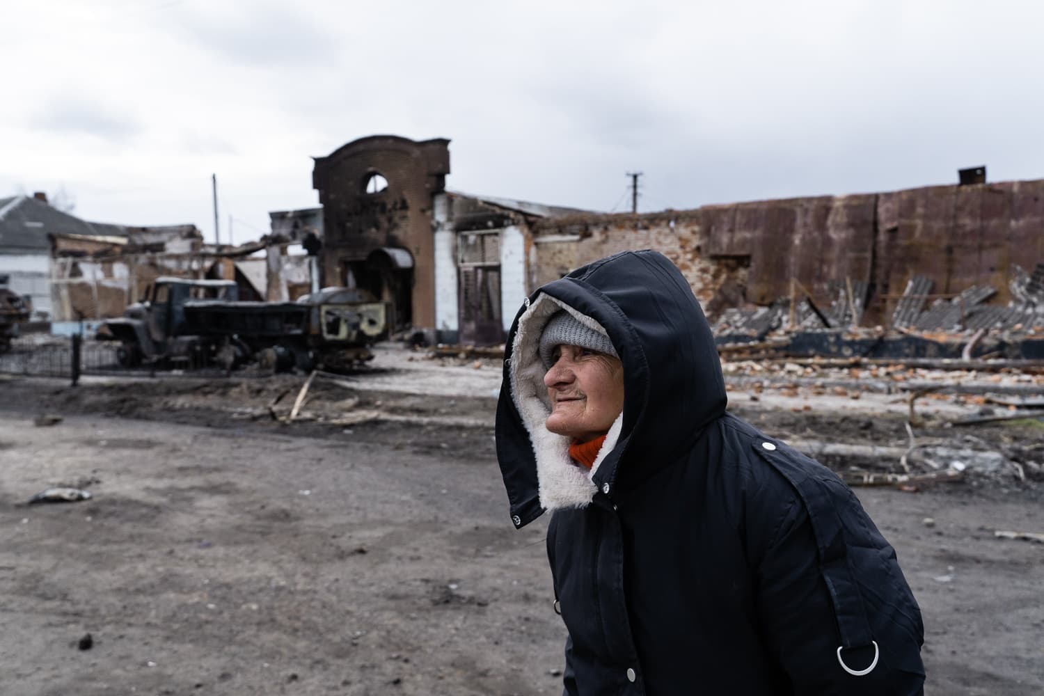 Женщина в капюшоне стоит на фоне разрушенных войной зданий и сгоревшего грузовика.