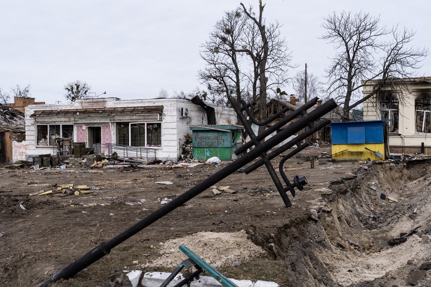 Разрушенный городской пейзаж: на переднем плане большая воронка от взрыва и обломки, наклонённый чёрный металлический столб. На заднем плане повреждённые одноэтажные здания, одно из которых — магазин, рядом киоск в цветах флага Украины. Надписи: "Валенский Скворец" (на киоске).