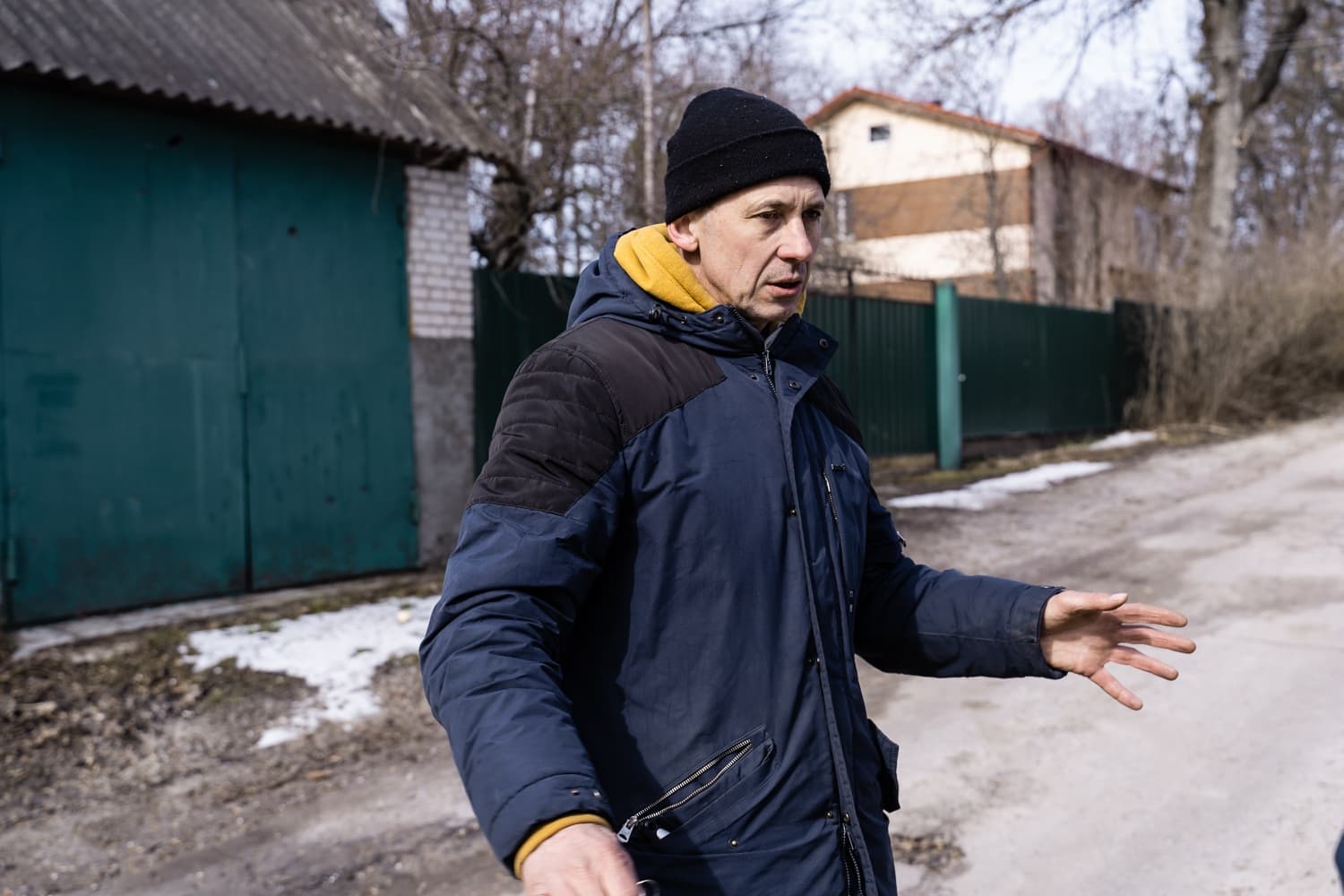 Мужчина в синей куртке и черной шапке жестикулирует на улице перед гаражом и жилым домом.