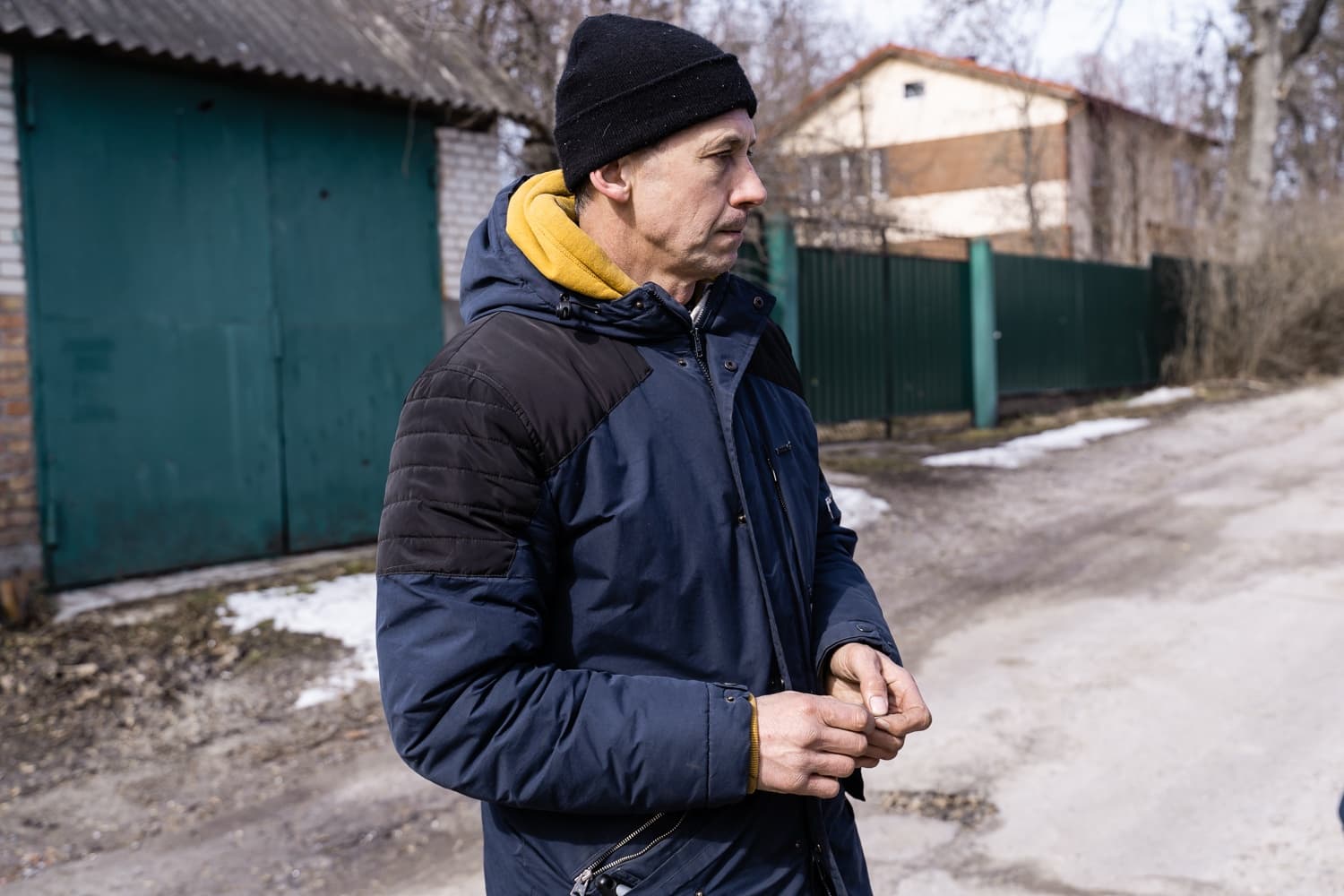 Мужчина в синей куртке, желтом худи и черной шапке стоит на улице на фоне гаража и частного дома.