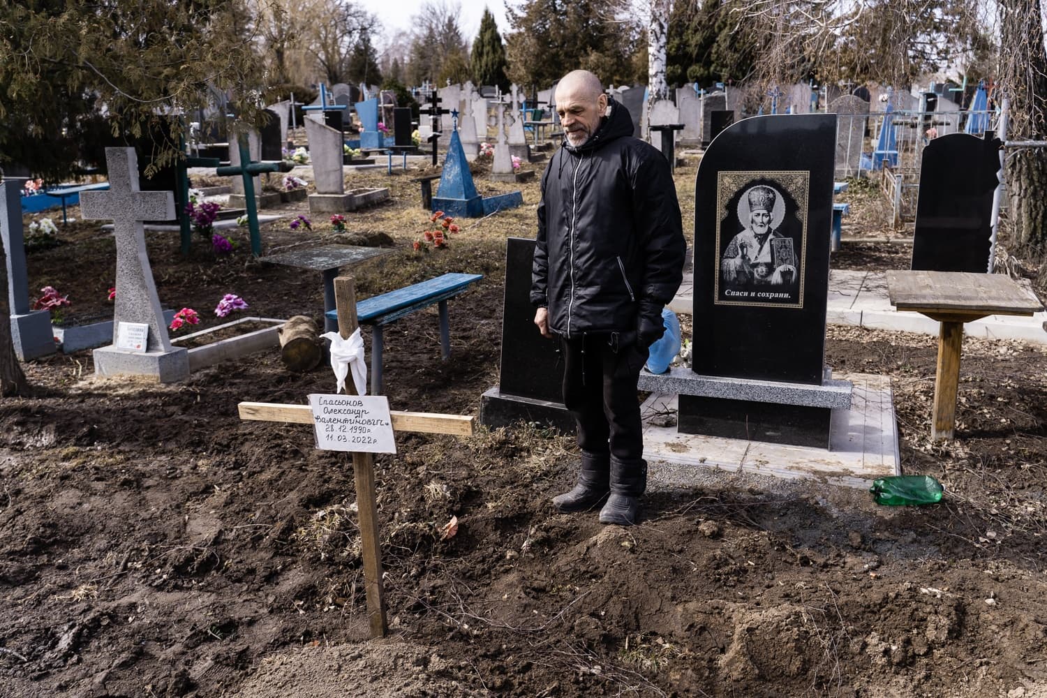 Мужчина в черной куртке стоит у свежей могилы на кладбище с временным деревянным крестом. На кресте написано: "Спасюков Александр Валентинович. 21.12.1990г. 14.03.2022г.". На соседнем надгробии: "Спаси и сохрани.".