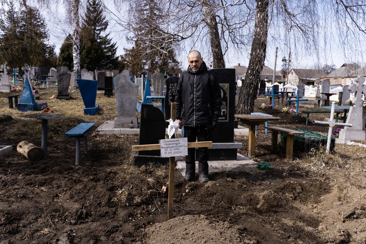 Мужчина стоит у свежей могилы на кладбище; на деревянном кресте надпись: "ЄЛАСЬКОВ ОЛЕКСІЙ ВАЛЕНТИНОВИЧ 28.12.1990 р. 11.03.2022 р.".