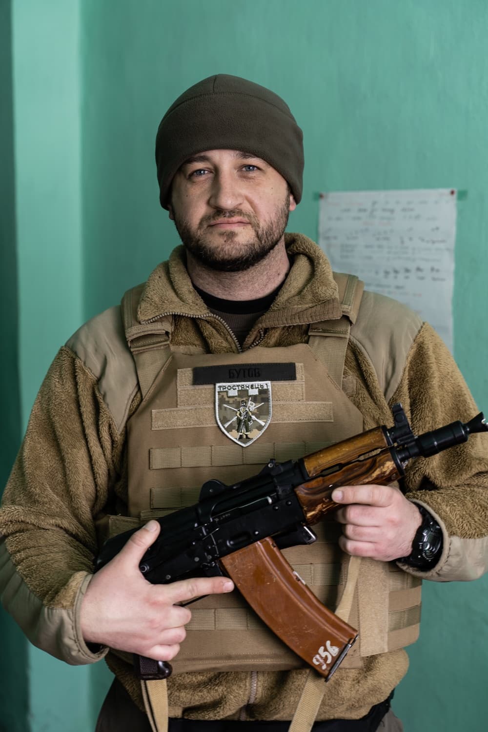 Военный в плитоноске и шапке держит автомат. На груди нашивки "БУГОВ" и "ТРОСТЯНЕЦЬ".